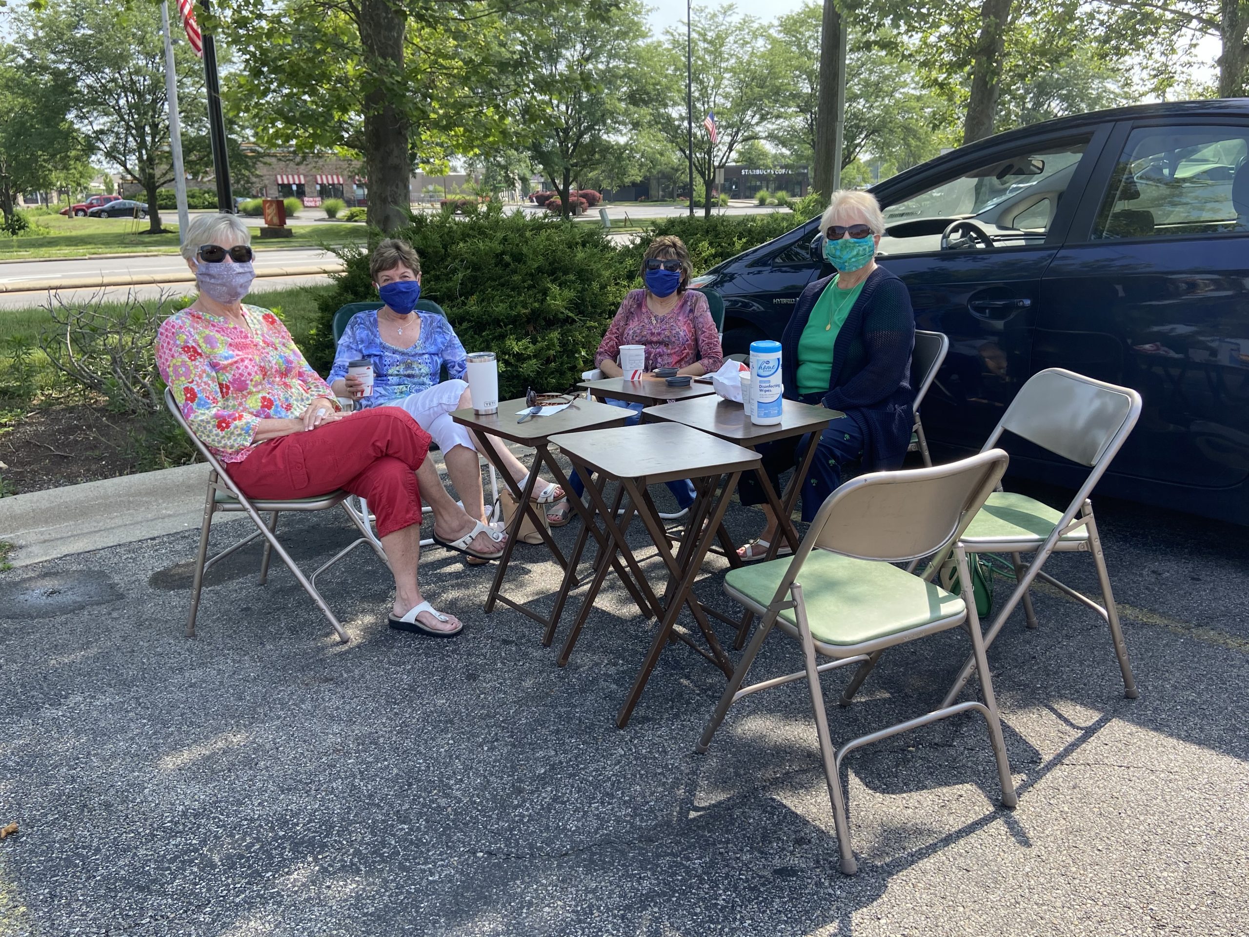 Four ladies are sitting around a make-shift table in a Chick-fil-A parking lot. They are wearing masks and have sanitizing wipes. They are enjoying spending time together.